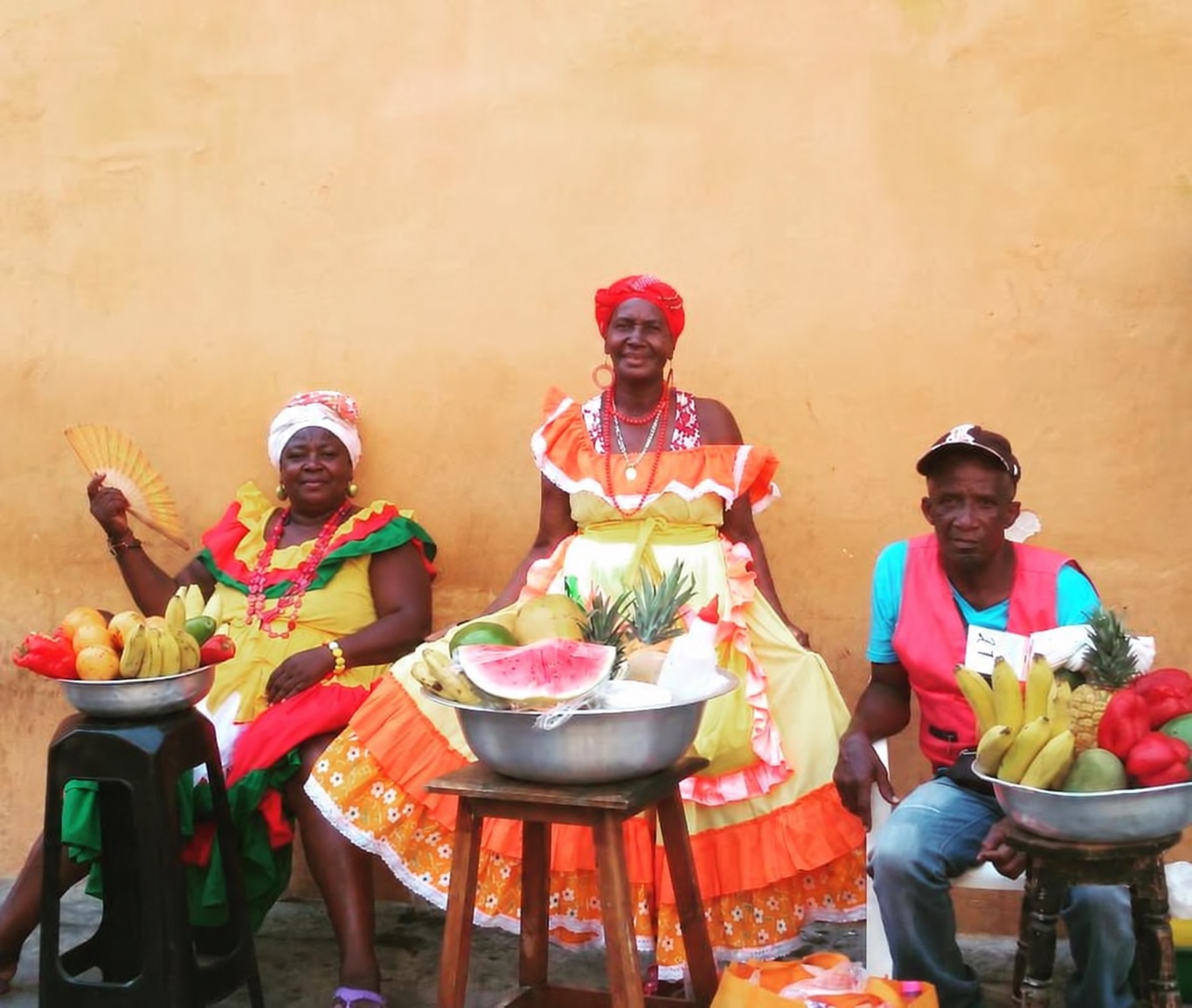 Encontro cultural na Colômbia: três pessoas com trajes coloridos junto a bancas de frutas. Foto de viagem de Anderson Nunes, com retoque digital.